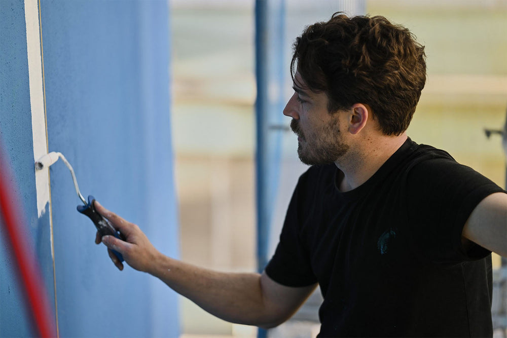 A man with dark hair and a beard uses a paint roller to paint a vertical white line on a blue wall. He is wearing a black T-shirt and appears focused on his work.