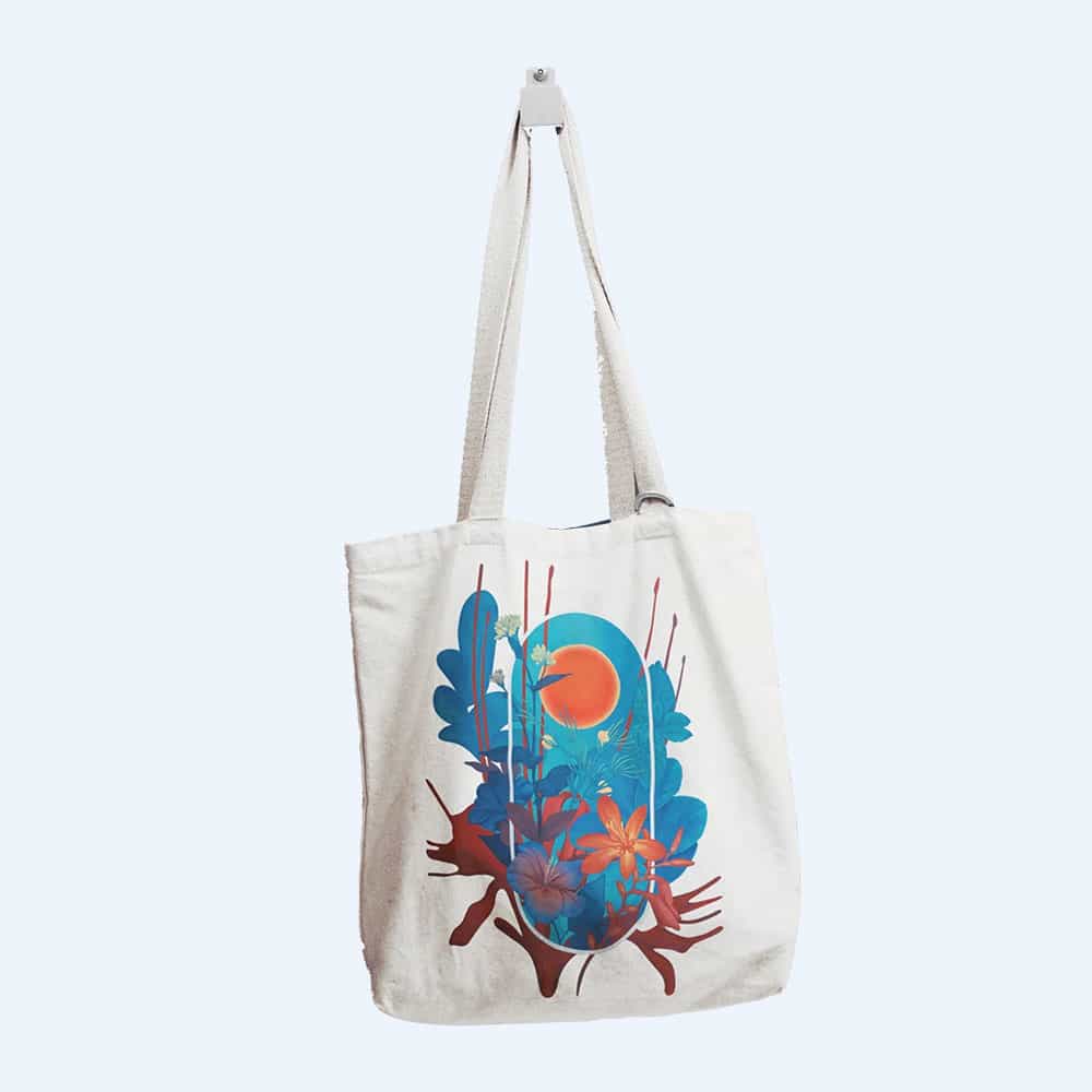 A cream-colored tote bag featuring a vibrant design of blue leaves, an orange sun, and red flowers, hanging on a white hook against a light background.