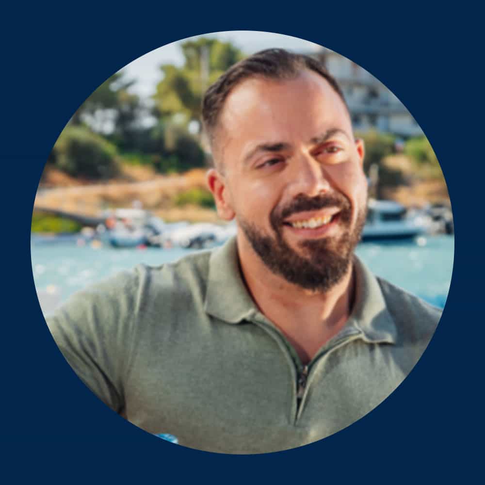 A man with a beard and short hair smiles while outdoors, wearing a light green polo shirt. The background shows water, boats, and trees under a clear sky.