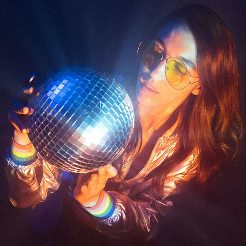 A woman wearing yellow-tinted sunglasses and a shiny jacket holds a reflective disco ball, surrounded by colorful light, creating a retro, party atmosphere.