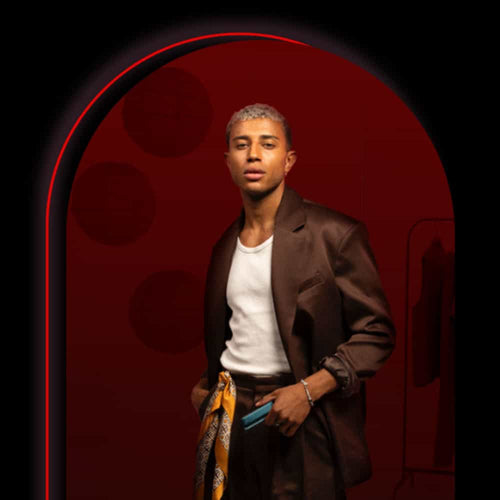 A stylish person with short, light hair stands confidently under a red-arched light. They wear a dark suit, white shirt, patterned scarf at the waist, and hold a phone, with clothes hanging in the dim background.