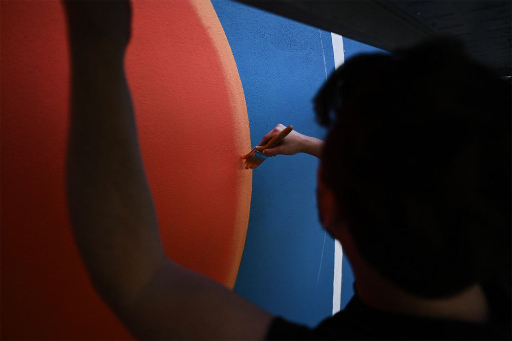 A person paints a border between a large orange-red circle and a blue background on a wall, using a small brush with careful attention to detail.
