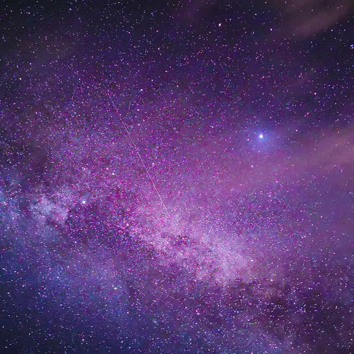 A vibrant night sky shows the Milky Way galaxy filled with countless stars, mostly in shades of purple and pink. A bright star stands out near the top right, and a faint line crosses diagonally through the stars.