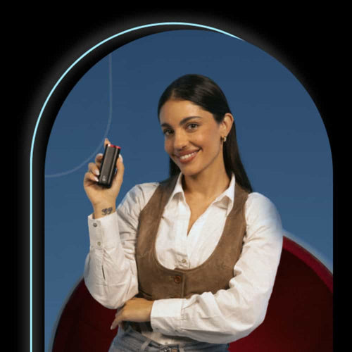 A woman with long dark hair, wearing a white shirt and brown vest, smiles while holding a small black device in her right hand against a blue background with a glowing arch.