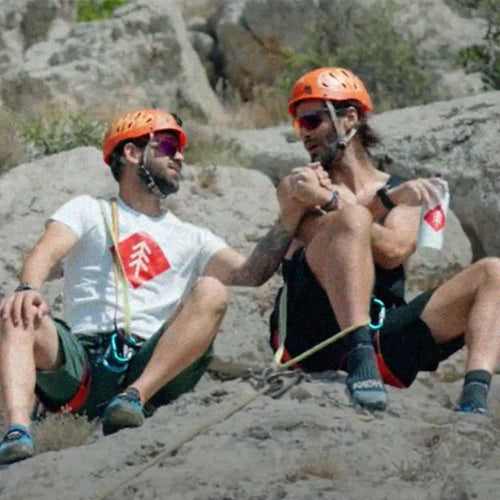 Two men wearing helmets and climbing gear sit on a rocky surface, smiling and holding hands, appearing to take a break while rock climbing outdoors.