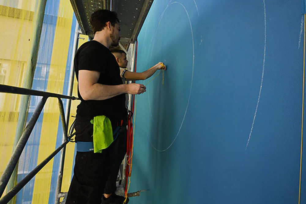 Two people stand on scaffolding, drawing large circular shapes with white chalk on a blue wall. One person is actively drawing while the other observes. Safety harnesses and tools are visible.