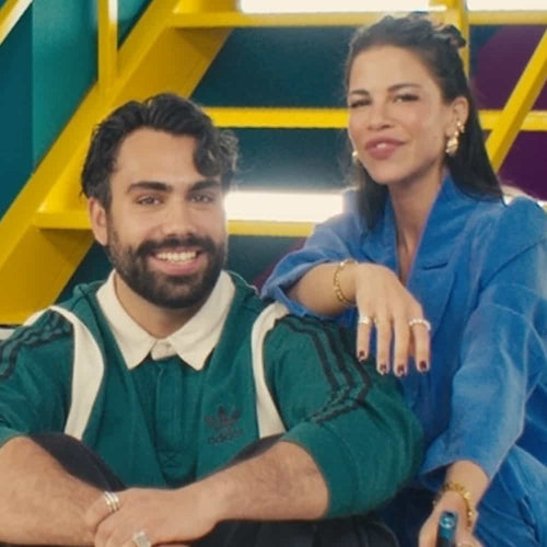 A man with dark hair and a beard, wearing a green shirt, sits next to a woman with long dark hair in a blue outfit. They are smiling and sitting in front of yellow stairs with colorful walls.