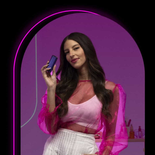 A woman with long dark hair, wearing a sheer pink top and white pants, stands in a purple-lit archway holding a small rectangular device. Shelves with colorful bottles are visible in the background.