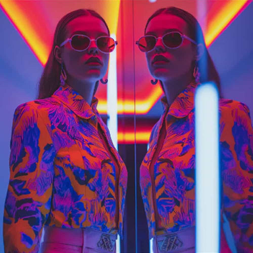 A woman in bold, colorful clothing and round sunglasses stands beside a mirror, creating a reflection. Neon pink, blue, and orange lights illuminate the scene with a vibrant, futuristic glow.