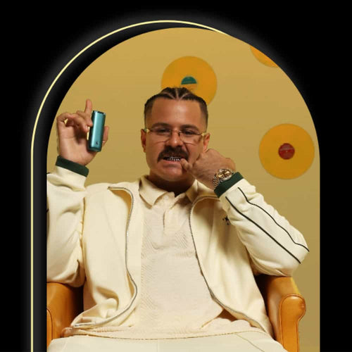 A man with slicked-back hair and glasses sits in a tan armchair, wearing a cream tracksuit. He holds a small blue device and gestures with his other hand. The background is yellow with circular decorations.
