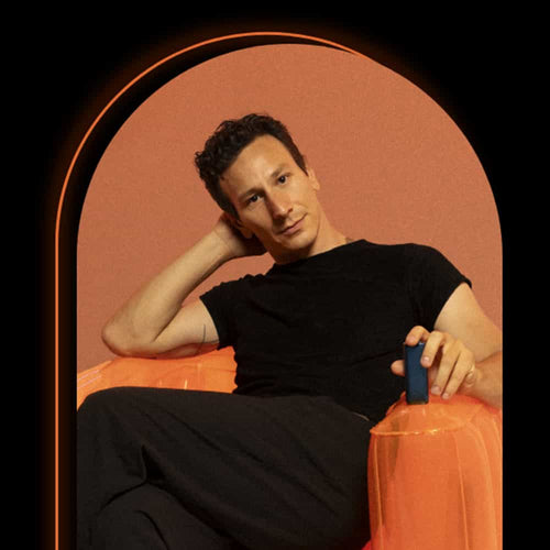 A person with short dark hair, wearing a black shirt and pants, sits casually on an orange inflatable chair against a peach-colored background, with one arm resting on the chair and the other behind their head.