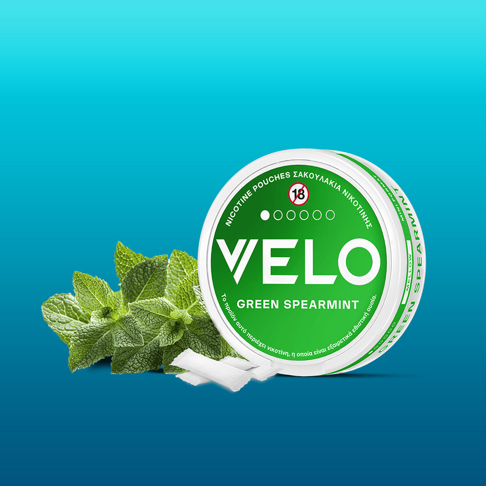 Ένα στρογγυλό δοχείο VELO FREE SAMPLE με πράσινες πινελιές εμφανίζεται δίπλα σε φρέσκα φύλλα δυόσμου και λευκά σακουλάκια σε μπλε διαβαθμισμένο φόντο - υποψήφιο προϊόν του έτους 2025 στην Ελλάδα.