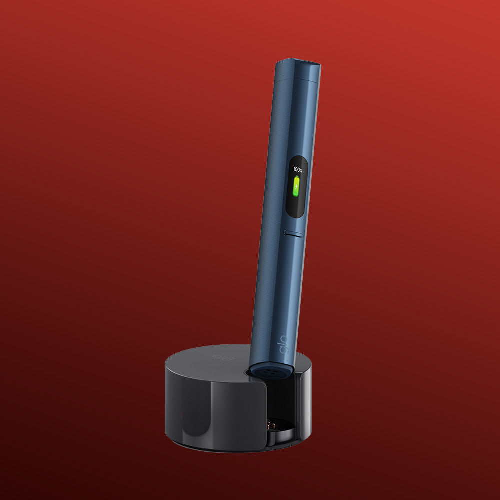 A slim, dark blue pen stands upright in a round black glo™ HILO Plus Charging Dock against a red gradient background. The device features a small digital screen showing battery status—always ready as one of your essential spare pens.